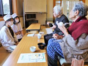 小学生2人の向かいに老人2人がおにぎりと汁を前に座っている様子。