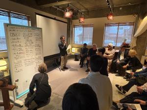 男性司会者が前に立ち、たくさん意見が書かれたホワイトボードの前に10人ほどの人が聞いている